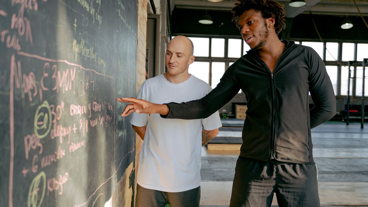 pranayama Two men planning a workout strategy on a blackboard in a modern gym setting.